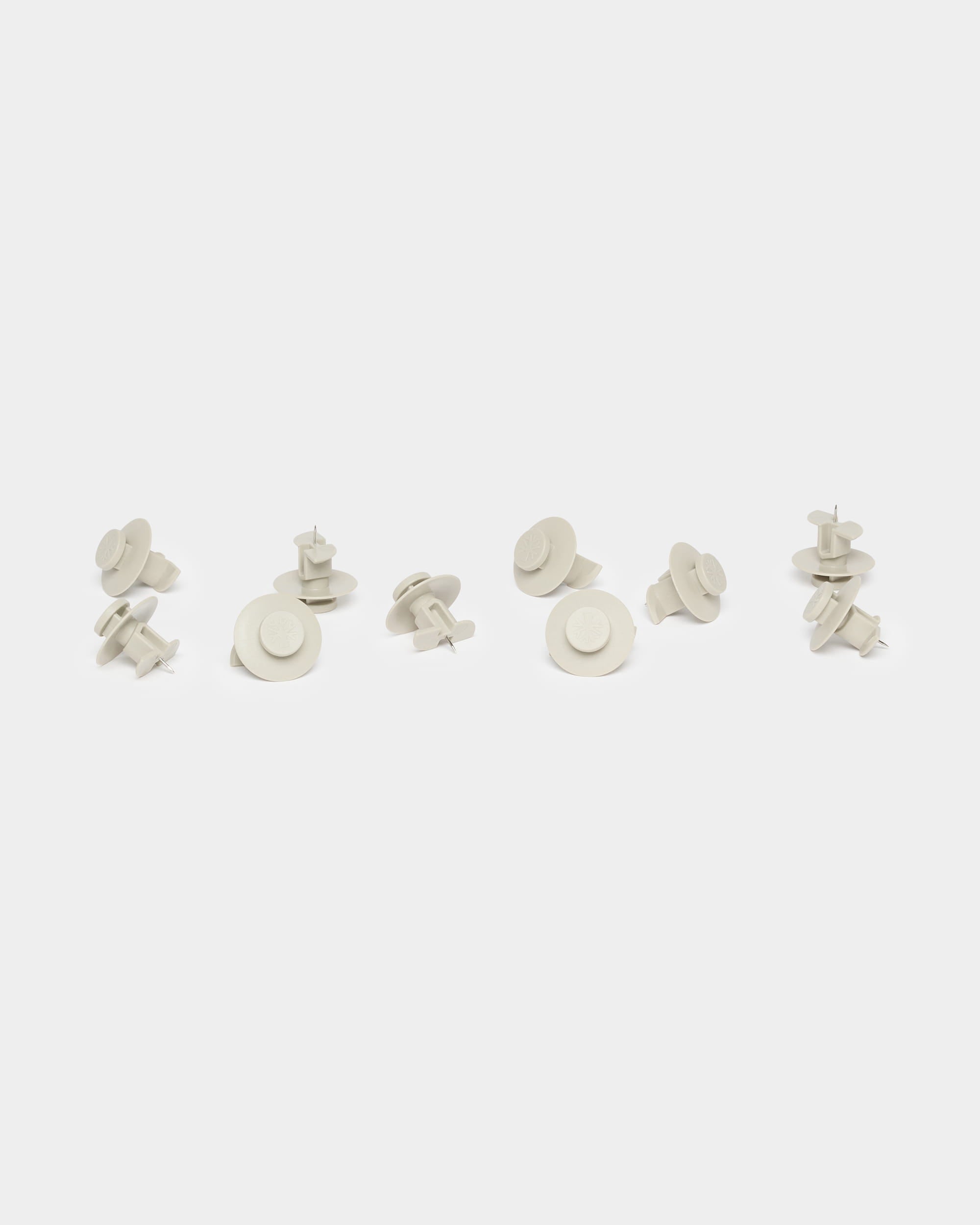 Dieci puntine da disegno Dupps color beige di WoodUpp, utilizzate come accessori per Akupanel, sono disposte in modo sparso su uno sfondo bianco semplice.